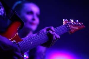 Elektrische Gitarre im Großformat samt Hand des Gitarristen, im Hintergrund eine Frau vermutliche Sängerin